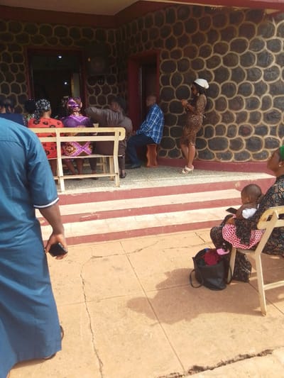 The scene shows a group of people gathered outside a building with a distinctive stone wall, potentially in Awka, Nigeria. A woman in a leopard-print dress and white head covering stands by the wall, holding a mobile phone. Seated nearby is a man in a blue patterned shirt. A bench holds several individuals, mostly women in patterned headscarves and clothing, with one man. In the foreground, a woman sits on a chair, holding a baby, with a black bag at her feet. A man in a blue robe stands in the left foreground, also holding a mobile phone. The scene is outdoors, on a paved area with steps leading up to the building.