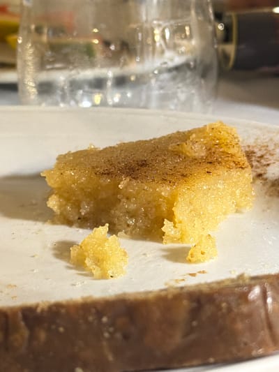 A close-up, overhead shot focuses on a golden-brown, rectangular piece of dessert, possibly a cake or pastry, dusted with cinnamon, on a white plate. A small, crumbled piece of the dessert sits to the left of the main piece. In the background, a blurred glass filled with water and ice is visible, reflecting light. The dessert appears moist and granular, with some sparkly sugar crystals. The setting suggests an indoor dining environment, likely a restaurant or cafe in the Sithonia Municipal Unit of Greece, given the food presentation and general ambiance. There are no people or discernible actions depicted in the image.