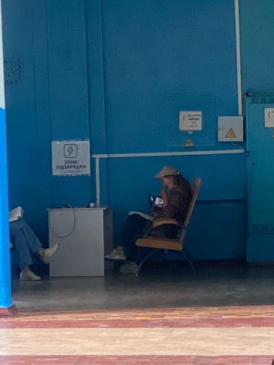 In Romodan, Ukraine, a person sits in a wooden chair, looking at a phone. The person is wearing a cap and a striped shirt. To the left, another person is visible, with legs crossed, holding a book. They are near a white cabinet with a charging sign above it. The environment appears to be indoors, with blue walls and a patterned floor. Electrical panels are visible on the wall in the background.