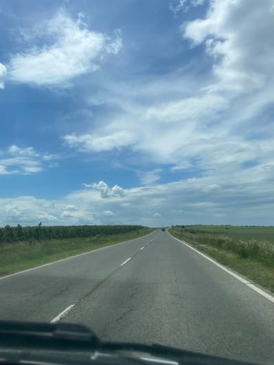 A slightly tilted view from inside a vehicle, capturing a straight, asphalt road stretching into the distance. The road is flanked by green vegetation on both sides. A blue sky with scattered white clouds dominates the upper portion of the frame. In the distance, a small, dark shape is visible on the road, presumably another vehicle. The scene may be located in Madara, Bulgaria.