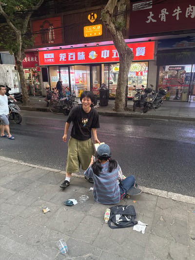 The image depicts an urban street scene in Changning District, China, likely at night, illuminated by artificial light. The wet asphalt street suggests recent precipitation. On the left, a man in a white t-shirt, grey shorts, and blue flip-flops stands near a black scooter, partially obscured.
In the foreground on the right, two individuals are interacting on a gray tiled sidewalk. A young woman with short dark hair, wearing a black t-shirt with a graphic design, olive-green wide-leg cropped pants, white socks, and black sneakers, stands smiling broadly. She holds a light-colored book or pamphlet in her left hand, extended towards a second individual. This second individual, seen from behind, is seated on the sidewalk with a black skateboard beneath them. They wear a dark blue baseball cap with "POLAR SKATE CO" visible on the back strap, a horizontally striped t-shirt (blue, light blue, white), and blue jeans, appearing focused downwards, possibly at the book or a device.
The sidewalk in the foreground contains scattered litter, including an empty plastic water bottle, crumpled paper, a crushed wrapper, a teal/blue JBL Bluetooth speaker with a white cloth, a light green plastic bottle, a black shoulder bag, and a white tissue.
In the midground, multiple scooters and a bicycle are parked along the sidewalk edge. Beyond, commercial buildings line the street. A prominent red facade features the Chinese characters "花百味" (Hua Bai Wei) and "中式快餐" (Chinese Fast Food), with a sign below indicating "早餐 5:30 营业" (Breakfast Open at 5:30). To the right, another large red sign displays "大骨牛肉" (Big Bone Beef). Above "花百味," a brown facade includes "万宏悦膳" (Wanhong Yueshan) and a yellow logo. Trees with thin trunks are interspersed along the sidewalk, and overhead utility lines are visible against the dark sky.