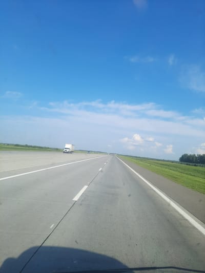 The image depicts a highway under a bright blue sky with scattered white clouds. A white truck is visible in the distance on the right lane of the highway. The highway is flanked by green grass on the right and a line of trees further in the distance. The perspective suggests the photo was taken from a vehicle moving along the highway. The location is identified as Pokrovka, Russia.
