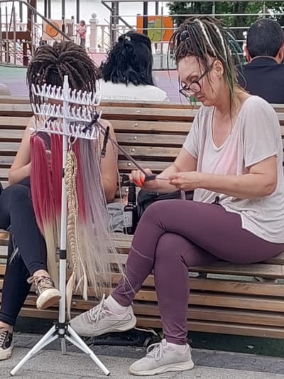 An outdoor street scene in Alikonovka, Russia depicts two women sitting on a wooden bench. The woman on the left has dark dreadlocks and is partially obscured by a white rack holding various colored hair extensions. The woman on the right, wearing glasses and a light-colored t-shirt with leggings, is actively braiding hair extensions. A tripod is positioned in front of them, and a black bottle is visible on the bench. In the background, a child is seen on a playground.