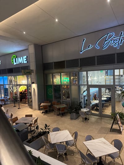 The image shows the exterior of two restaurants, "Le Bistro" and "Lime," at night. "Le Bistro" has a cursive, illuminated sign above its glass-fronted entrance. Patrons are visible inside. Outdoor seating is arranged with tables and blue and white chairs. Further down, "Lime" is visible, with its green and white sign. The restaurant also has a glass facade. Tables and chairs are set up outside.