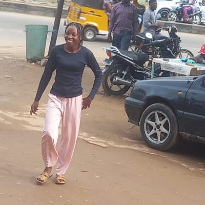 A woman is standing on a street in Dadi Riba, Nigeria, smiling and facing the camera. She is wearing a long-sleeved dark blue shirt, light pink pants, and light brown sandals. A black car is parked to her right, and several motorcycles are parked behind the car. A yellow auto rickshaw is visible in the background. Other people are in the vicinity. The environment appears to be a bustling urban street.