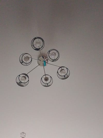A modern, multi-arm chandelier is mounted on a white ceiling in a room in Yanino-1, Russia. The chandelier has a central chrome fixture with six arms extending outwards. Each arm holds a light bulb enclosed in a cylindrical shade with an open, circular metal frame. The room appears to be empty, with only the chandelier and a portion of the ceiling visible. The lighting is soft, suggesting ambient or artificial light.