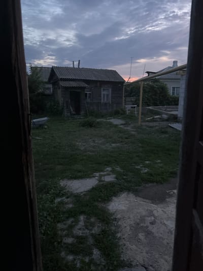 This image depicts a dimly lit outdoor scene, likely taken at dusk, in a rural or suburban area of Novoanninskiy, Russia. In the foreground, a dark, wooden doorway or window frame is visible, partially obscuring the view.
The central focus is a small, weathered wooden structure with a corrugated roof. To its left, another similar structure with a corrugated roof is partially visible. A wheelbarrow, light gray in color, sits on the grassy ground to the left of the wooden house. The ground is covered in patchy grass and concrete or stone elements.
In the background, other buildings with roofs are visible, and a yellow metal pipe or railing runs horizontally across the mid-ground. The sky is cloudy with shades of purple, gray, and hints of orange from what could be a setting sun. The overall impression is one of a simple, possibly rustic, residential setting.