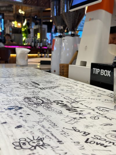 The image shows a close-up of a white wall covered in black marker drawings and handwritten text. The perspective is from a low angle, looking up at the wall which appears to be part of a counter or table. Many of the drawings are simple cartoons, including a spider-like creature, a cat face, and a peace sign. The handwriting is varied, with some words clearly legible like "Loves", "Some", "Song", and "TEL". Some more complex phrases are also present, such as "Gerrntte!" and "So amazing how much the world size". There are also some numbers and dates visible.
To the right of the wall, a white and orange point-of-sale (POS) terminal is visible, with a black "TIP BOX" attached to it. Further in the background, out of focus, are glimpses of a busy indoor setting with people, lights, and what appears to be a bar area with bottles. The lighting suggests it could be daytime or evening. The overall impression is a casual and interactive environment where patrons are encouraged to leave their mark. The presence of the POS terminal and tip box indicates a commercial establishment, likely a cafe, bar, or restaurant. The text "Baan Chaweng, Thailand" is provided as location context, suggesting this establishment is in that area.