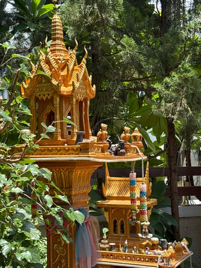 The image captures an outdoor scene at Baan Karnkanok 23 in Thailand, focusing on a miniature golden shrine or spirit house. The shrine is ornately decorated with intricate carvings and tiered roofs, typical of Thai religious architecture. It is adorned with colorful fabric ties and flower garlands. Several small figurines, including lion-like creatures and human figures, are visible on and around the shrine. The shrine is situated amidst lush greenery, with large, broad leaves and coniferous branches framing the structure. Sunlight filters through the foliage, casting shadows and highlighting the golden sheen of the shrine. The overall impression is one of a serene, spiritual space integrated within a natural environment.