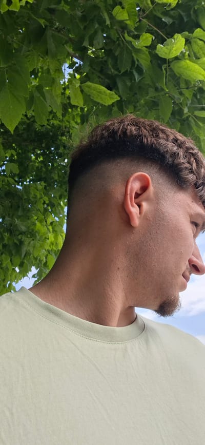 A low-angle, close-up shot captures the side profile of a person from the neck to the upper head, set against a backdrop of lush green leaves and glimpses of a blue sky. The person, appearing to be male, is seen looking downwards or away from the camera. They wear a light-colored, possibly light green or beige, t-shirt, with the ribbed collar and shoulder visible.

Key details include a faded haircut on the side of the head, and what appears to be braided or cornrowed hair on the visible portion of the head towards the right. There is stubble on the cheek and jawline, and a small goatee under the lower lip. The ear is clearly visible. The background is dominated by a dense canopy of bright green leaves, with patches of clear blue sky peeking through, indicating a sunny daytime. The image provides no specific activity other than the person being present outdoors in this natural setting. No text is visible in the media file. The location context for this image is Strazhitsa, Bulgaria.