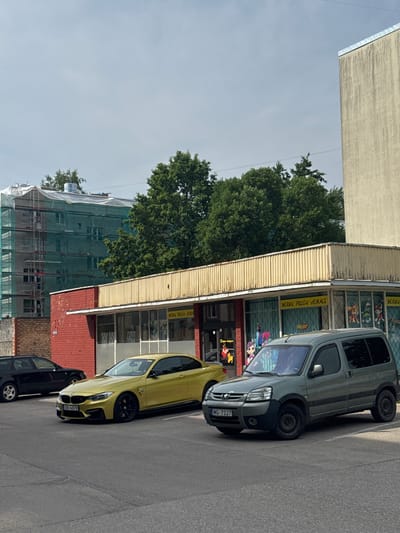 The image shows a street scene in Olaine, Latvia. A grey van and a yellow car are parked on the side of the road, in front of a building with a yellow corrugated metal awning. The building has large windows with colorful cartoon characters displayed. One window has text that reads "BĒRNU PREČU VEIKALS". To the left of the building, a large green tree is visible, and further to the left, a building is covered in green scaffolding and netting, suggesting construction or renovation. The sky is overcast, indicating daytime. The overall impression is of a residential or commercial area on a slightly cloudy day.