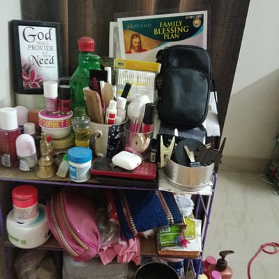 The image depicts a three-tiered purple shelving unit overloaded with miscellaneous items. On the top shelf are numerous cosmetic and personal care products, including bottles, jars (one labeled "SORBET SU"), a small golden bell, and containers of various sizes. A framed print that reads "God shall provide all your need" stands at the back, along with a "Jesus Calls Family Blessing Plan" booklet, and a green dinosaur figurine. Also present are mugs holding combs, brushes, and other items. On the middle shelf, a red cellular phone, a white earpod case, and a metal container holding keys and other small items are visible. The bottom shelf contains a few larger containers of cream and a pink makeup bag. The image was taken indoors and appears to be in a domestic setting, likely within the city of Bengaluru, India.