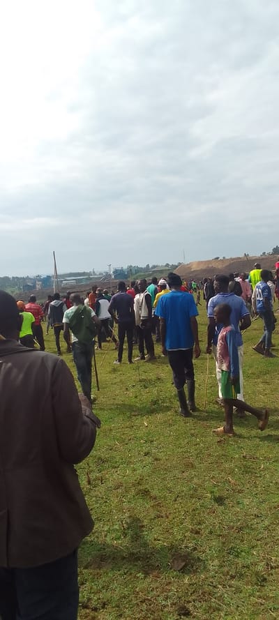 The image shows a group of people standing in a grassy field. There are at least 30 people in the image, most of them are men and some are children. Most of the people are wearing casual clothing. The people are looking in different directions, and some are holding sticks. The scene is taking place in an open field with a small hill in the background. The sky is cloudy. The location is in Lubao, Kenya, at coordinates (latitude: 0.386, longitude: 34.824).