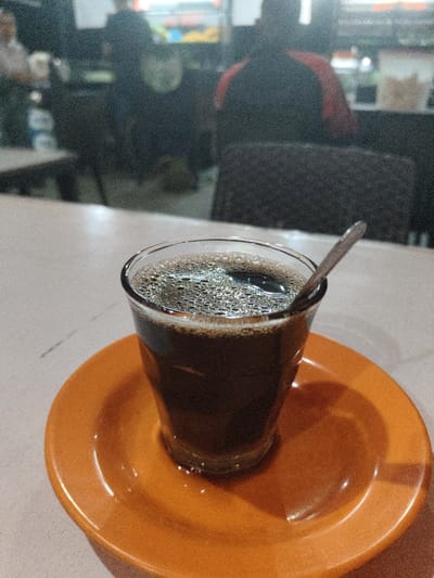A clear glass containing a dark, bubbly liquid, consistent with coffee, occupies the foreground, positioned on a bright orange saucer. A silver spoon rests submerged in the liquid. The glass and saucer are situated on a light-colored, possibly off-white or beige, tabletop. The background is out of focus, revealing the interior of an establishment, likely an eatery or cafe, located in the city of Rheng, Indonesia. Several blurred human figures are visible in the background. One prominent figure, seen from behind, is seated in a dark chair, wearing a dark garment with a distinct orange or red stripe on the upper arm/shoulder area. Other indistinct figures and partial views of tables and chairs are also present. The scene is illuminated by ambient lighting, contributing to a slight graininess in the image. There are no clear actions or interactions depicted due to the blur.