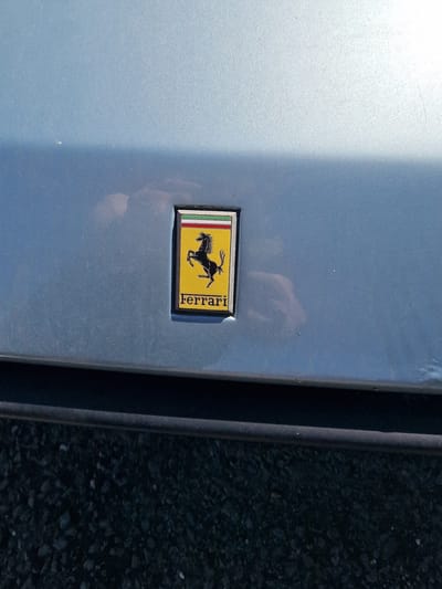 The image presents a close-up, eye-level view of a light blue or silver-grey car body panel, likely a door or fender, illuminated by bright daylight. A rectangular Ferrari shield emblem is centrally placed on the car's surface.
The emblem features the iconic black "Cavallino Rampante" (prancing horse) on a vivid yellow background. To the left of the horse, three vertical stripes in green, white, and red are visible, representing the Italian flag. The brand name "Ferrari" is written vertically in black capital letters along the right side of the yellow background. The emblem has a dark, possibly black, border.
The car's paintwork appears slightly textured or dusty, with a bright reflection of light, likely sunlight, on the upper left portion of the panel. To the right of the car panel, a dark vertical line or trim separates it from an out-of-focus background of dark asphalt or gravel, indicating an outdoor setting. No people are visible, and no specific activity is taking place. The scene suggests a clear, sunny day. The only visible text is "Ferrari" on the emblem.