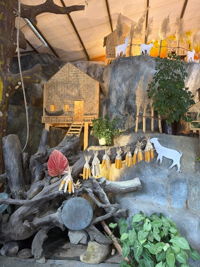The image shows a detailed diorama depicting a rural Vietnamese village scene, possibly set in Khanh Vinh. The diorama is built against a rough, grey, rocky backdrop, suggesting a mountainous or hilly terrain. In the upper section, two small, thatched-roof houses are visible, nestled into the rocks. Several white, stylized goat figures are placed on the rocks, with some standing near the houses and one in the middle distance. Tall, feathery plants, resembling reeds or pampas grass, are scattered throughout the scene, adding a natural element.
In the mid-ground, a larger, more detailed thatched-roof house is prominently displayed. It has a wooden staircase leading up to a raised platform with a small balcony. The house features small windows and a doorway, illuminated from within, suggesting it is occupied. In front of this house, a large, gnarled tree trunk and root system forms a base. A bright red, fan-shaped leaf is placed on the roots.
Below the mid-ground house, a series of dried corn cobs, tied in bundles, hang from a piece of wood, creating a decorative element. A white, stylized goat figure is positioned to the right of these corn bundles, appearing to interact with them. Further down, at the base of the diorama, is a collection of weathered logs and rocks. A large, dark, circular object, possibly a stone disc or a shallow bowl, rests on some rocks amidst the logs. Lush green plants with heart-shaped leaves grow at the lower right corner of the scene, adding a vibrant touch of nature. The overall impression is a carefully constructed miniature landscape that evokes a sense of rustic simplicity and traditional village life.