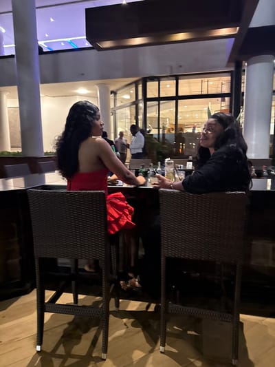 The image displays two women seated at a high table or bar within an indoor commercial establishment in Itirin, Nigeria, during evening hours. The woman on the left, with long, dark, wavy hair, wears a red strapless dress featuring a ruffled skirt. She is seated on a dark woven bar stool, positioned facing slightly right, engaged in conversation with her left arm resting on the dark, reflective tabletop. Opposite her, the woman on the right, also with long, dark, wavy hair, wears a dark top and occupies a matching bar stool. She faces left, looking towards the first woman with a slight smile, and holds a clear glass containing a beverage and straw. The tabletop holds additional clear glasses, small bottles (including a distinct dark green one), and other indistinct items. In the background, through a large glass partition with dark frames, several blurred male figures are visible in an adjacent, brightly lit interior space. One man in a white collared shirt and another in a light-colored top are discernible. White cylindrical columns and overhead lighting, including some with a bluish hue, are also present in the background. The floor beneath the bar stools is light-colored.