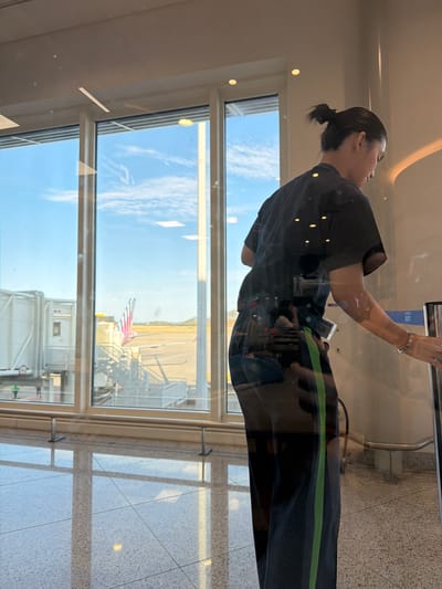 A person wearing a black shirt and black pants with a green stripe is standing in front of a large window at Athens International Airport in Spata-Loutsa Municipal Unit, Greece. Through the window, part of an airplane with an American flag on the tail is visible. The person appears to be interacting with something out of view, possibly a scanner or a gate. The surrounding environment includes tiled flooring and the infrastructure of the airport, like jet bridges.