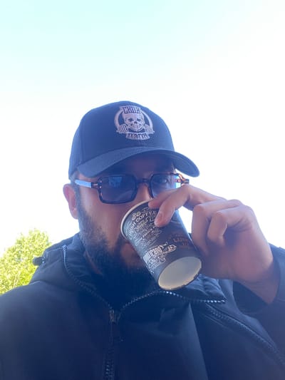 A person, wearing a black baseball cap with a skull logo and brown-rimmed sunglasses, drinks from a black paper coffee cup. The person has a beard and is wearing a black jacket with a zipper. The background consists of a bright, clear sky and a green tree. The photo was taken outdoors.