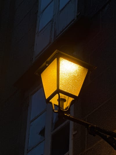 The image shows a close-up, low-angle shot of a vintage-style, yellow-lit street lamp against a dark, textured building facade. The lamp has a square, frosted glass enclosure that diffuses the warm light, and it is mounted on a black metal arm. The building appears to be constructed of stone or concrete and has several window frames visible, some of which show cracked or broken glass. The overall atmosphere is dark and moody, suggesting it is nighttime. The architectural style and the presence of a stone building are consistent with the historic city of Santiago de Compostela, Spain, though a specific location is not identifiable from the image alone. There are no people or animals present.