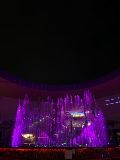 A nighttime scene depicts a large, illuminated water fountain display, characteristic of an amusement or entertainment park. The location is consistent with Batu Night Spectacular (BNS) in Batu, Indonesia.

The primary subject is a multi-jet fountain, projecting numerous streams of water vertically, predominantly illuminated in a vibrant magenta-purple hue. Green light beams are visible traversing horizontally through the water plumes. The fountain structure rests on a raised, textured base, accented with red-orange lighting.

In the background, behind the fountain, a building features an illuminated sign clearly displaying "AQUA STATION" in blue and yellow lettering, with smaller text visible below it. To the left, a portion of a large, ornate sign reads "HOUSE OF TERROR." The upper section of the image shows a dark sky transitioning into a curved architectural element, possibly a ceiling or facade, illuminated by purple light, suggesting a large indoor or semi-enclosed venue. No individuals are visible in the frame. The overall environment is characterized by artificial lighting against a dark backdrop.