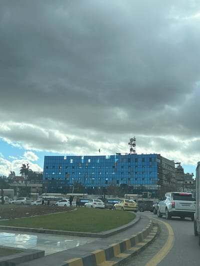 An overhead-level view depicts a busy urban street scene in Al-Muhajerin Municipality, Syria, under a predominantly overcast sky with grey clouds, interspersed with patches of lighter clouds and blue sky. The focal point is a large, multi-story building with a facade composed entirely of reflective blue glass panels, featuring numerous square windows, some appearing open or internally lit. A dark flag is flown from a pole on the roof, near the left side of the blue building, while a prominent, multi-tiered communication tower occupies the right side of its roof.
In the foreground, a paved road features a black and yellow striped curb bordering a central median of green grass, transitioning to an area of dry earth. To the far left foreground, a wet surface or puddle reflects the sky. Traffic is visible on the road, including several sedans of varying colors (white, yellow) and a white SUV positioned prominently on the right. Pedestrians are observed near the median on the left side of the street, walking or standing. Additional buildings, including one with a red roof and visible palm trees, line the background on the left, while other urban structures extend to the right of the central blue building.