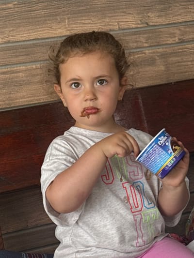 A young girl with curly brown hair and light brown eyes sits on a wooden bench, holding a cup of ice cream with a spoon. Chocolate is smeared around her mouth. She is wearing a grey t-shirt with colorful lettering and pink leggings. The background consists of a wooden wall. The image appears to be taken outdoors, possibly in the city of Rusokastro, Bulgaria.