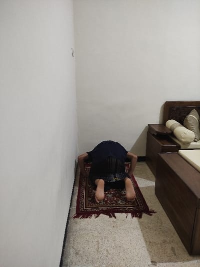 The image shows a man kneeling in prayer on a red prayer rug, his head bowed in front of a plain white wall, in a room located at coordinates (-7.828, 112.701) in Kecamatan Lawang, Indonesia. The man is wearing a dark long-sleeved shirt and dark pants. The prayer rug has a fringe border. There is a partially visible wooden bed frame with cushions to the right of the man, and a matching wooden footboard or nightstand with a dark surface in the lower right corner. The flooring is a light-colored speckled tile, and the walls are white, with a light switch visible in the upper left corner of the image. The room is otherwise empty.