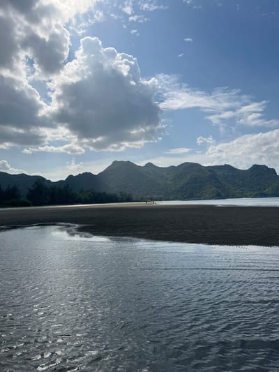 The image depicts a coastal scene in Ban Hup Ta Khot, Thailand. In the foreground, shallow water with ripples reflects the sky. A dark sandbar extends into the water, with a thin strip of water on its further side. Beyond the sandbar, a line of trees separates the beach from a range of lush green, mountainous terrain. The sky is a mixture of blue sky and dramatic, sunlit clouds, with the sun partially obscured. Two small figures, appearing to be people, are visible walking on the sandbar in the mid-ground. The overall scene is natural and serene, dominated by water, sand, vegetation, and mountains under a dynamic sky.