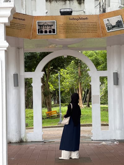This is a vertical, medium shot photograph taken outdoors in Ratsada, Thailand. The scene depicts a woman wearing a black hijab, a black top, and a dark blue skirt, standing on a brick pathway. She is carrying a light-colored crossbody bag. The woman is partially obscuring a display board that contains text and images.
The display board is positioned to the left of the frame and is partially visible. It features two images that appear to be old photographs of buildings, resembling stamps. The text on the board is in Thai script and appears to be historical information. Visible text includes "โรงเรียนภูเก็ตไทยหัว" (Phuket Thai Hua School), and "คมนาคมขนส่ง" (Transportation and Communications). The content suggests that the display is about the history of a school or a local area.
Behind the woman and the display board, there is a lush green garden with trees. The architecture in the foreground features white pillars and arched openings, suggesting a covered walkway or a historical building. The lighting indicates it is daytime, with soft shadows, suggesting it might be late afternoon or an overcast day. The overall impression is one of historical and cultural exploration in a garden setting.