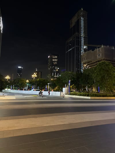This is a night-time, eye-level, full shot of a modern urban scene in Huangpu District, China. The image is dominated by the dark night sky, illuminated by the lights of several tall buildings. In the foreground, a paved plaza with a geometric pattern of light and dark rectangular tiles stretches across the frame. Several people are visible. On the left side of the plaza, a person on a scooter is traveling from left to right. A second person, also on a scooter, is slightly further to the right and ahead of the first. To the right of these individuals, a person in a red jacket walks towards the left. Beyond the plaza, a white barrier runs horizontally, separating the paved area from a strip of green foliage and trees. Several street lamps with multiple globes are positioned along this barrier, casting a warm yellow light. Behind the trees and lamps, more buildings rise into the night sky. The most prominent of these is a very tall skyscraper, partially under construction, with its skeletal structure and scaffolding visible. Another tall building to its left is fully illuminated, with many windows glowing. A crane is visible near the construction site. The overall impression is one of a bustling urban environment at night, with ongoing development.
