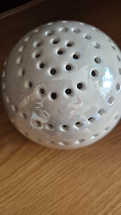 The image shows a close-up of a white, spherical object with numerous small, dark holes covering its surface. The object appears to be made of ceramic and has a slightly iridescent sheen. It is resting on a wooden surface with visible grain. The lighting suggests an indoor setting, possibly a home environment, and there are reflections on the object that indicate the presence of other objects or light sources. There is no indication of any specific location in Miltenberg, Germany.