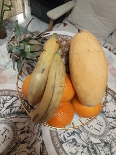 A high-angle close-up shot reveals a gold wire fruit basket filled with assorted fruits, sitting atop a patterned round table. The fruit selection includes two ripening bananas, a large mango, several tangerines, and the leafy crown of a pineapple. The location appears to be indoors. The visible text on the table reads "RUB YOU," "EXIF TO," "'S FEET AMERICAN STYLE," "Y," "SOR AND RU," "SU," "AID," and "LEATHERWEAR MAT BUDON LE." The surrounding area has a neutral-toned fabric backdrop, possibly a cushion or part of furniture. The scene includes a bamboo plant in a vase and a white chair in the background. The fruit basket appears to be placed on a table in Baan Chaloklum, Thailand.