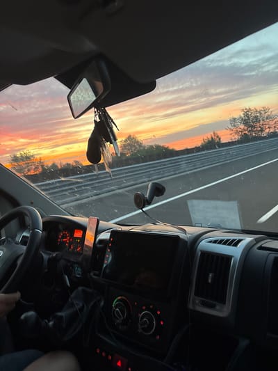 The image depicts the interior view from the passenger side of a vehicle driving on a highway at sunset. The dashboard is visible, featuring a steering wheel, instrument cluster, a central infotainment screen, and air vents. A set of keys hangs from the rearview mirror. The windshield offers a view of the highway, with guardrails and trees lining the side, silhouetted against a colorful sky with hues of orange, pink, and yellow. There is no visible indication of the specific location being Świętoniowa, Poland, within the image itself.