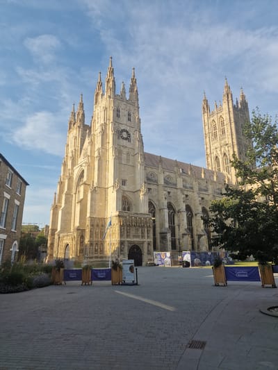 This media file displays a low-angle, vertical shot of Canterbury Cathedral, a grand Gothic structure. The cathedral is illuminated by the warm, golden light of either early morning or late afternoon, casting long shadows on the paved courtyard. The sky above is a clear, pale blue with wispy white clouds.
In the foreground, the cobblestone pavement leads towards the cathedral, with planters containing greenery and several tall, blue banners lining the path. These banners display the text "Canterbury Cathedral" in white lettering. A large tree with dark green foliage partially obscures the upper portions of the cathedral's spires and towers in the top right of the frame.
The cathedral's intricate facade is visible, featuring numerous arched windows, decorative carvings, and sharp spires reaching towards the sky. A prominent clock face is visible on one of the towers. In the lower portion of the image, a brick building with arched windows is partially visible, suggesting an urban setting. The overall scene evokes a sense of awe and historical grandeur.