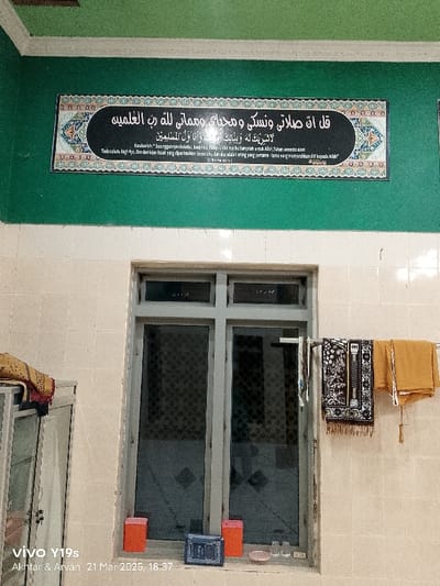 The scene is a room with white tiled walls and a green tiled ceiling. On the wall above a large window is a framed picture of a religious text in Arabic. The window is made of glass panes and has a metal frame. There are two red boxes on the windowsill below the window, and several small glasses are to the right of the boxes. There are two prayer rugs hanging from a metal bar outside the window. The scene is located in the Kecamatan Kroya district of Cilacap Regency, Indonesia. The coordinates of the location are -6.423 latitude and 107.996 longitude.