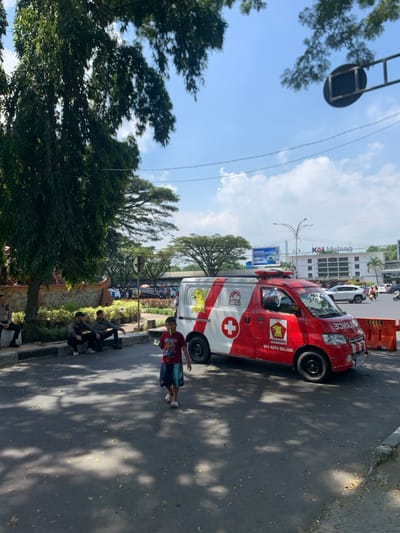 The image shows a red and white ambulance parked on a paved street in Malang, Indonesia, at the coordinates -7.978, 112.637. The ambulance is marked with a white cross, the words "DPC Kota Malang," and a red eagle with the word "REHINDA" underneath. A young boy in a red shirt and blue shorts stands on the sidewalk, facing the camera. To the left of the ambulance, three adult males are seated on a curb, dressed in dark clothing, looking in different directions. One of the men has his right leg extended. The street is lined with low green bushes. A large green tree with a brown trunk can be seen on the left side of the image. A tall, multi-story building with a white facade and a billboard sign that says "KAI Malang" can be seen in the distance. The sky is a bright blue with a few white clouds.