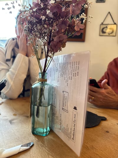 This image depicts an indoor scene within a cafe or restaurant in Ambleside, United Kingdom. The foreground features a light blue-green glass vase containing a bouquet of dried flowers, predominantly maroon-colored hydrangea-like blooms and small white flowers, tied with twine. To the right of the vase, a clear plastic menu stand displays various beverages and food items, including "Breakfast loose leaf Tea £3.25" and "Soft Drinks" with prices in GBP. The items rest on a light brown wooden table with visible grain, where a metal spoon is partially visible, wrapped in a white napkin.

In the mid-ground on the left, a person with fair skin, wearing a white ribbed sweater over a striped garment, is seated with their hands near their face, partially obscured by the flowers and strong backlight from a window behind them. On the right, another person, wearing a reddish-brown long-sleeved shirt, is holding a dark smartphone with their left hand; a gold ring is visible on their ring finger. A dark, possibly slate, coaster is partially visible beneath the menu stand. The background shows a light-colored wall with framed decor, including a small, gold-framed piece depicting a reflective C-shape design.