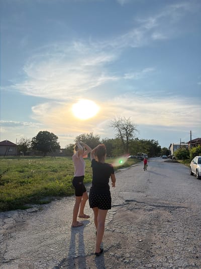 The image depicts a rural scene in Dolni Dabnik, Bulgaria, during what appears to be late afternoon or early evening. Two young women are walking away from the camera on a paved road. The woman on the left wears a pink t-shirt and black shorts, while the woman on the right is dressed in a black t-shirt and polka-dot shorts. Both have their arms raised, touching their hair. In the distance, a child rides a bicycle down the road. The road is lined with houses, trees, and parked cars. The sun is low in the sky, casting a warm glow and creating long shadows. The sky is a clear blue with wispy clouds. To the left of the road, there is a grassy field with scattered dandelions. The overall atmosphere is serene and peaceful.