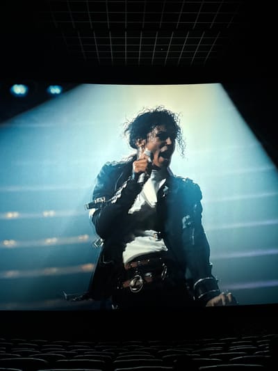 The image depicts Michael Jackson singing into a microphone on stage. He is wearing a black jacket over a white shirt, and his signature curly hair is visible. The background appears to be a brightly lit stage, with vertical light beams. The image is slightly tilted, and the surroundings appear dark, possibly the interior of a venue. The location in Taihe District, China is not visible in the image.