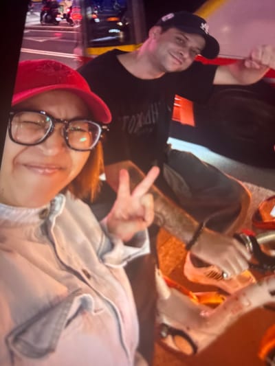 This media file captures a close-up, low-angle shot of two individuals, likely friends, enjoying themselves in what appears to be an outdoor urban setting at night.

In the foreground, a person wearing a red baseball cap and glasses is seen from the chest up, lying down and looking upwards with a slight smile. Their hair is reddish-brown. They are wearing a light-colored denim jacket.

To the left and slightly behind them, another person is visible from the shoulders up. They are wearing a black t-shirt and a black baseball cap with a white logo. Their head is turned towards the camera, and they appear to be smiling or smirking, with their right hand raised in a thumbs-up gesture.

The background is blurred but suggests a vibrant, possibly festive atmosphere, with hints of colorful lights and structures, consistent with a night scene in a city like Taipei, Taiwan. The presence of what looks like streetlights and possibly traffic lights implies an urban environment.

One individual's arm and hand are visible, making a peace sign. There is some visible text on the t-shirt of the person on the left, but it is partially obscured and difficult to read clearly. The overall mood seems relaxed and happy, suggesting a casual outing or celebration. The lighting indicates it's nighttime.