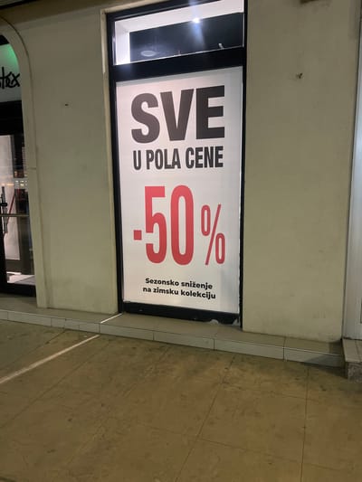 The image displays a commercial storefront at night in Smederevska Palanka Municipality, Serbia. A large white poster covers the main glass panel of the entrance. The poster features dark gray capital letters "SVE" (ALL) at the top, followed by "U POLA CENE" (AT HALF PRICE) in smaller dark gray capital letters. A prominent red "-50%" is centered beneath, with smaller dark gray text at the bottom reading "Sezonsko sniženje na zimsku kolekciju" (Seasonal discount on winter collection). The storefront glass is framed in black. To the immediate left, a portion of another glass door with a visible metallic handle is present, adjacent to a light-colored arched architectural detail. To the right of the main entrance, a flat, light-colored wall segment is visible. The ground consists of light-colored square pavement tiles, with a low tiled threshold separating the exterior from the building. A linear light reflection diagonally crosses the pavement in the lower-left quadrant. No individuals or active interactions are depicted.