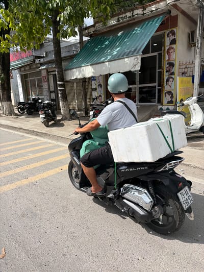 A street scene in Cam Ranh, Vietnam, is captured on a bright, sunny day with strong sunlight casting shadows. The main subject is a person, seen from behind, wearing a light blue helmet, grey t-shirt, and dark shorts or pants, operating a black Honda Airblade scooter. A large white styrofoam box is prominently secured to the back of the scooter with green straps.
The setting appears to be a commercial street, with buildings lining the right side of the road. One building features a green-striped awning and a yellow sign with red lettering. Visible Vietnamese text on this sign includes "PHƯƠNG" and "HỘI TÓC XINH," which translates to "Beautiful Hair Salon." Another partially visible sign shows "BÉ TA." Several headshot images of different hairstyles are displayed below the main sign, indicating a barbershop or hair salon.
Further down the street, another white scooter is parked. A tree with lush green foliage is visible on the left side of the frame, partially obscuring other buildings. The road surface is asphalt, with distinct yellow double lines painted on it. The scooter's license plate, "79-AA 590.52," is clearly visible. The overall impression is one of typical daily activity in a Vietnamese urban environment.