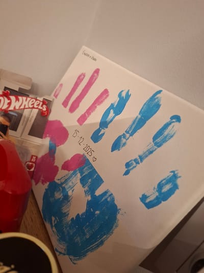 A close-up shot of a canvas displaying painted handprints in blue and pink. The handprints are arranged with blue prints above and pink prints below. Text "Evelina + Ivan" is written in the top left corner of the canvas, and "15-12-2025" with a heart is written in the middle. The canvas sits on a surface partially obscured by other objects, including a red plastic container and what appears to be a Hot Wheels toy box. The location is unknown.
