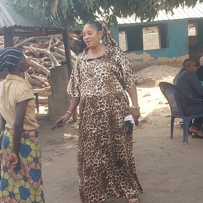 An outdoor scene in Assai Sabongida, Nigeria, features three individuals on unpaved ground under bright daylight. In the left foreground, a woman stands with her back partially to the camera, wearing a light brown short-sleeved top and a skirt patterned with blue, yellow, and orange swirls, her hands clasped behind her. She faces an adult woman positioned centrally. This central woman, facing towards the left, wears a full-length leopard print kaftan, large light blue earrings, and a matching patterned head wrap. She holds a black smartphone in her left hand and a white cloth in her right, displaying gold-colored bangles on her left wrist and a gold-colored watch on her right wrist. In the right mid-ground, an adult male is partially visible, seated in a blue plastic chair. The background elements include a large pile of cut logs or firewood stacked under a corrugated metal roof structure on the left. To the right, a partially constructed building with blue-painted walls and open window frames is visible, surrounded by mounds of earth. Green foliage from overhead trees is present at the top of the frame.