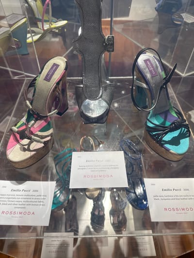 This is a close-up shot of a retail display of women's shoes, likely in a boutique or museum exhibit. The shoes are primarily high-heeled sandals and wedges, presented in clear plastic cases. The style of the shoes is varied, with some featuring intricate strap work, others with bold geometric patterns, and at least one pair in a textured, possibly snakeskin-effect material. The brand "EMILIO PUCCI" is visible on labels attached to several pairs of shoes, indicating they are part of a collection from this designer. Text on the labels also includes "ROSSIMODA" and "VENEZIA 1947," suggesting the display is associated with this establishment in Venice, Italy. One label specifies "Emilio Pucci 2006" and another "Emilio Pucci 2007," providing a timeframe for the showcased items. The lighting suggests an indoor setting, possibly a well-lit shop or exhibition space. The overall impression is one of a curated collection of designer footwear.
