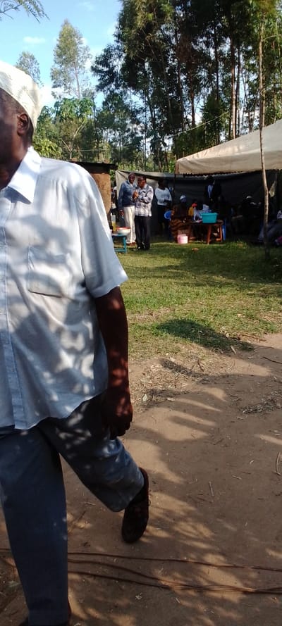 The image shows a man walking on a dirt path in Kipkarren, Kenya. He is wearing a white shirt, grey pants, and dark brown shoes. The man is walking away from the camera towards the right side of the frame.  The background shows a group of people gathered under a white tent in a grassy area. Two men are standing together in the center of the image, one wearing a light blue shirt and the other a dark shirt. Several other people are sitting or standing around the tent. Trees surround the scene.