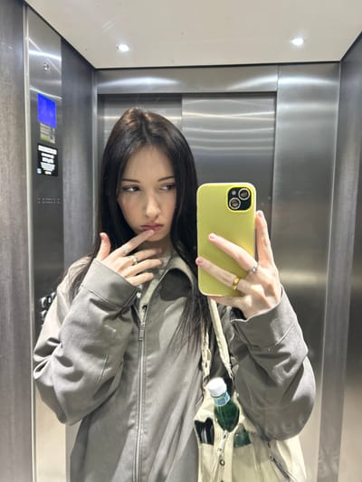 A young female is depicted taking a self-portrait reflection inside a metallic elevator car in Cachan, France. She has long, dark brown hair, a light complexion, and wears a grey zippered jacket over a lighter-colored top. Her left hand, adorned with silver- and gold-colored rings, is raised with the index finger touching her lower lip, while her right hand holds a smartphone in a bright yellow case, positioned to capture the image. Pink nail polish is visible on her right hand. A light-colored fabric tote bag hangs from her left shoulder, containing a green glass bottle with a white label. The elevator interior features reflective brushed metal walls, a control panel with blue illuminated elements on the left, and recessed spotlights on the ceiling. The subject's gaze is directed slightly to her left, with a pensive expression.