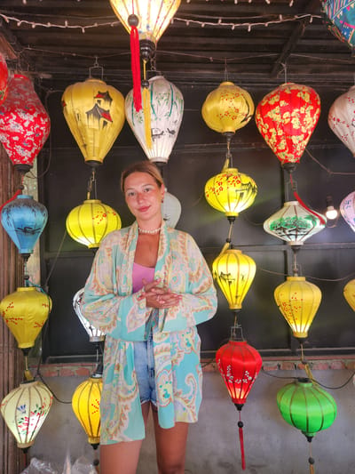 A woman stands in front of a wall adorned with numerous colorful, illuminated silk lanterns. She is wearing a light blue and beige patterned kimono-style robe over a pink tank top and denim shorts. Her hands are clasped in front of her. The lanterns are in various shapes and colors, including red, yellow, blue, green, and white, many featuring painted designs of landscapes and flora. The scene appears to be an outdoor market or shop in Hội An, Vietnam, a city known for its lantern-making tradition. The lanterns are hung at different heights and are illuminated from within, casting a warm glow.