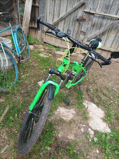 The image shows two bicycles in what appears to be a rural setting. A bright green mountain bike is in the foreground, angled to the right. It has knobby tires, a suspension fork, and a black seat with a green accent. Visible branding on the bike includes "SHIMANO" and "GREEN SPORT". A blue bicycle is in the background, to the left, with a rear rack and a fender. Both bicycles are parked on a dirt ground with patchy grass and some scattered stones and debris. Behind the bicycles is a weathered wooden fence and a portion of a wooden structure, likely a shed or outbuilding. The overall environment suggests an outdoor area, possibly a yard or an alleyway, with a rustic and somewhat unkempt appearance. The location is identified as Shurma, Russia. There are no people present in the image.